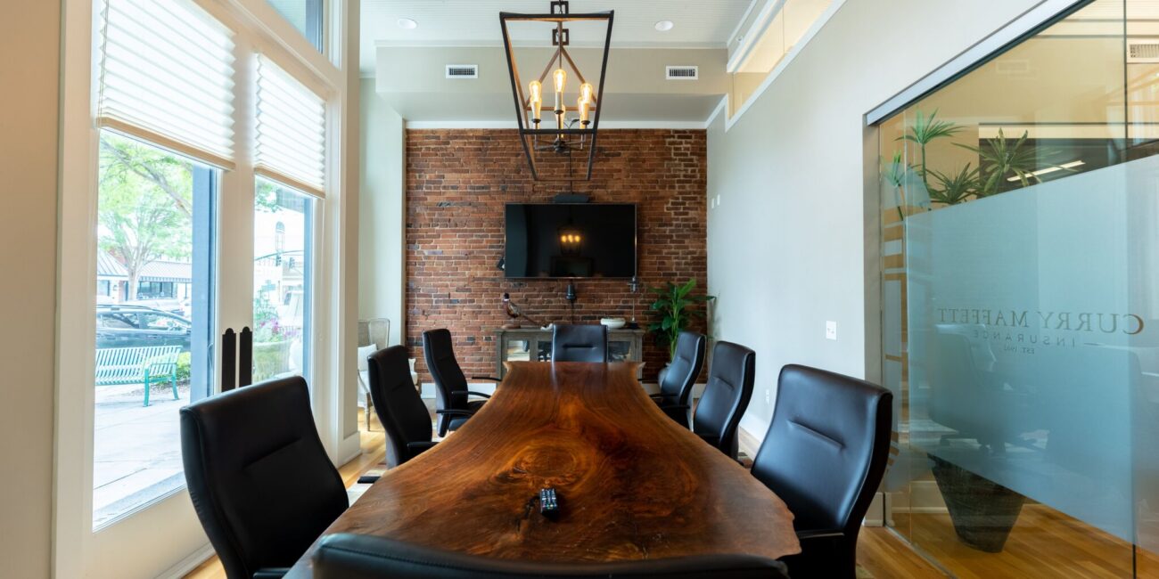 A modern conference room with a live-edge wood table, black chairs, brick wall, TV, large windows with blinds, and glass wall with logo.