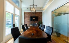 A modern conference room with a live-edge wood table, black chairs, brick wall, TV, large windows with blinds, and glass wall with logo.