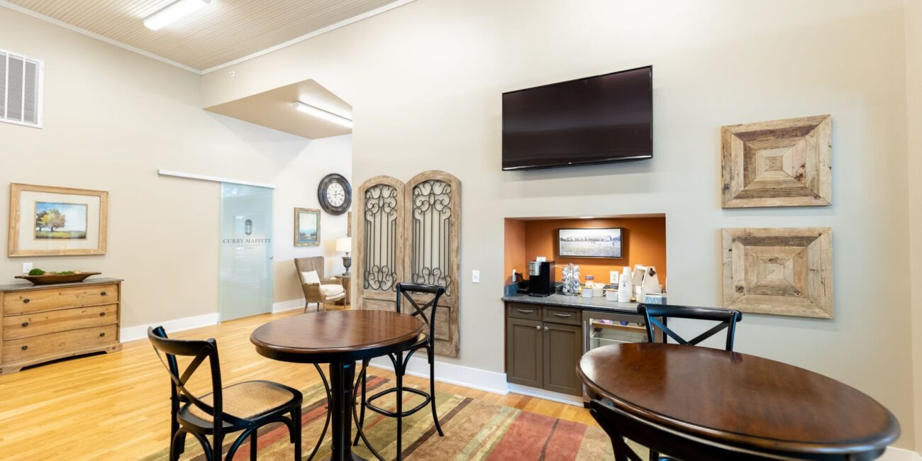 A bright lounge with round wooden tables, black chairs, a TV, coffee station, framed art on beige walls, and a colorful rug.
