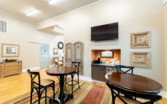 A bright lounge with round wooden tables, black chairs, a TV, coffee station, framed art on beige walls, and a colorful rug.