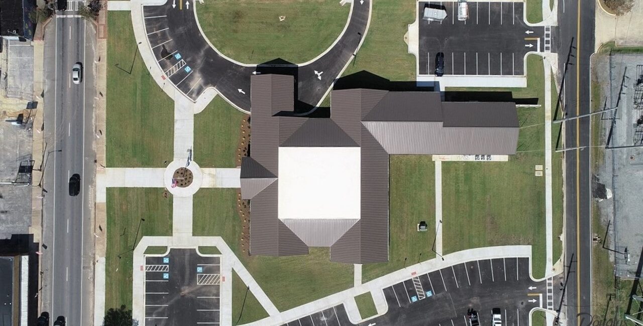 Aerial view of a modern, large building with a cross-shaped roof, green lawns, sidewalks, parking lots, and bordering urban streets.