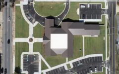 Aerial view of a modern, large building with a cross-shaped roof, green lawns, sidewalks, parking lots, and bordering urban streets.