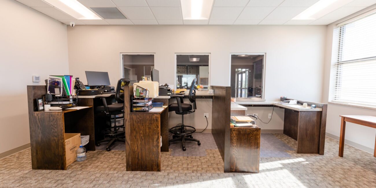 Three wooden desks with chairs in a row face a glass window, each with computers and supplies. Sunlight streams through the right window.
