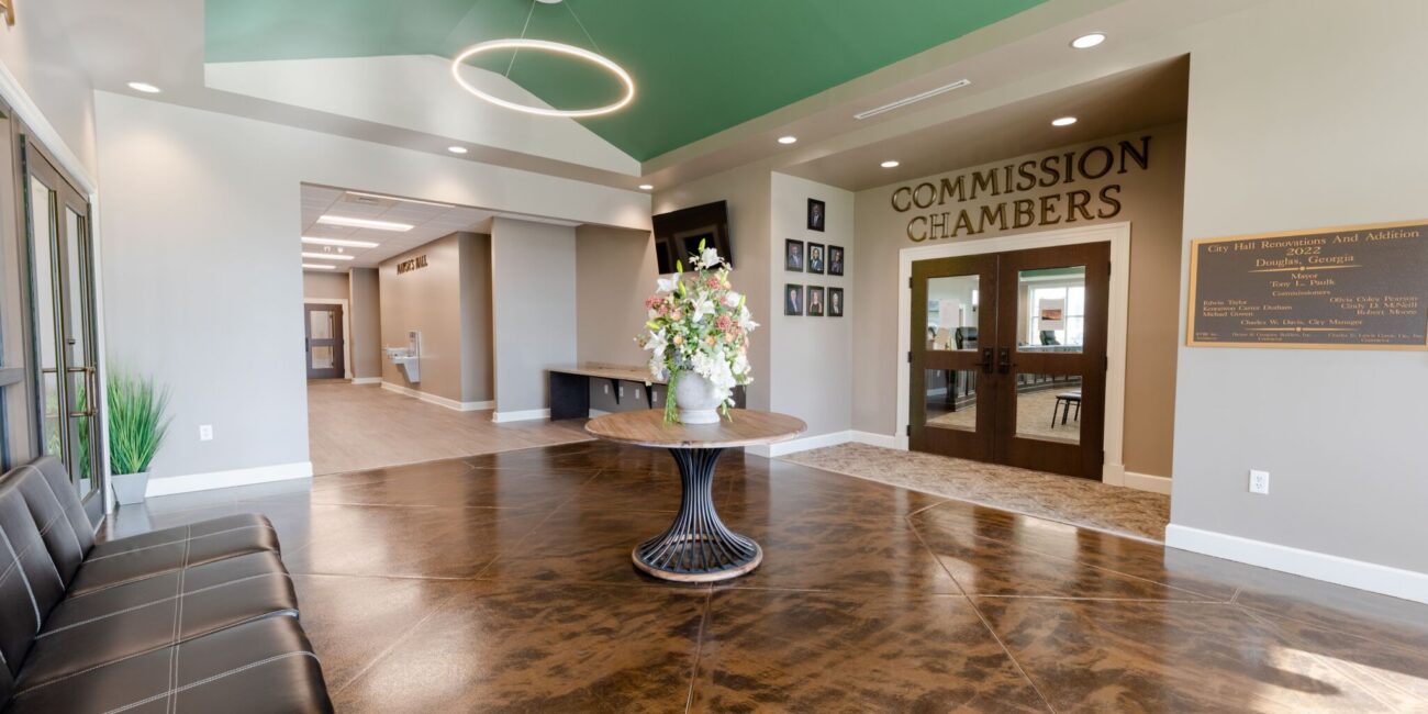 Spacious, modern lobby with a dark leather bench, round table with flowers, shiny patterned floor, and double doors labeled Commission Chambers under a green ceiling with circular light fixtures.