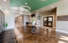 Spacious, modern lobby with a dark leather bench, round table with flowers, shiny patterned floor, and double doors labeled Commission Chambers under a green ceiling with circular light fixtures.