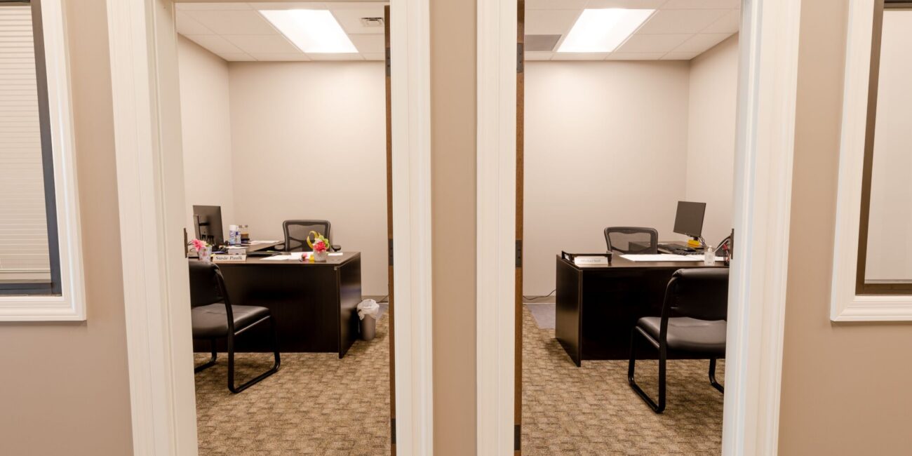 Two adjacent small offices with desks, computers, chairs, and supplies. Beige walls, carpeted floors, seen from the hallway.