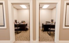 Two adjacent small offices with desks, computers, chairs, and supplies. Beige walls, carpeted floors, seen from the hallway.