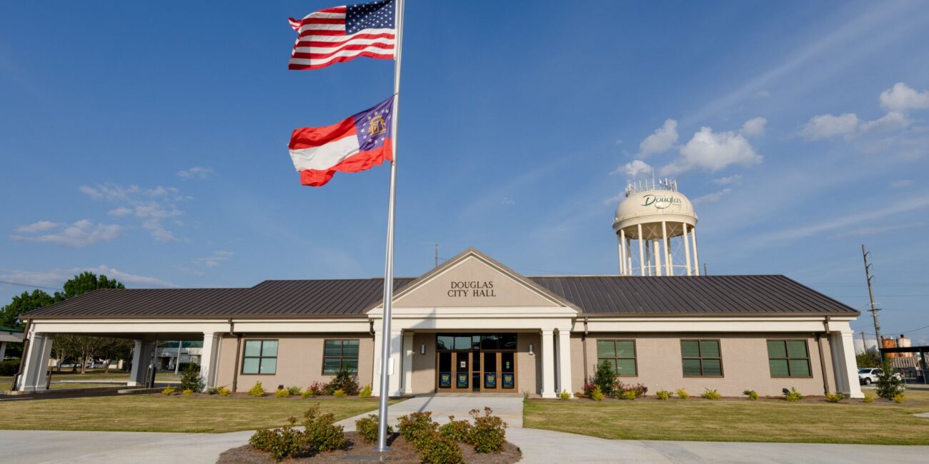 A one-story building labeled Douglas City Hall with American and Georgia state flags and a water tower labeled Douglas under a blue sky.