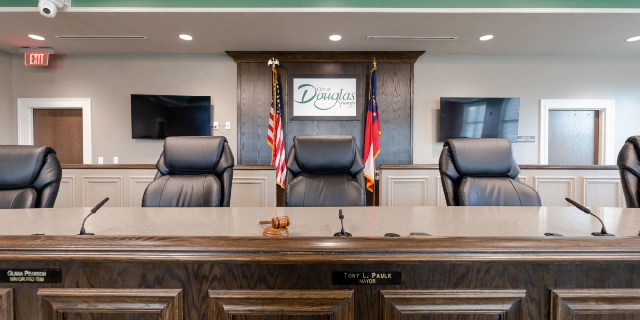 A council chamber with empty black leather chairs, a wooden desk with nameplates and gavel, two flags, Douglas sign, TVs, and doors.