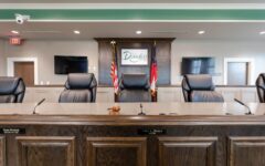 A council chamber with empty black leather chairs, a wooden desk with nameplates and gavel, two flags, Douglas sign, TVs, and doors.