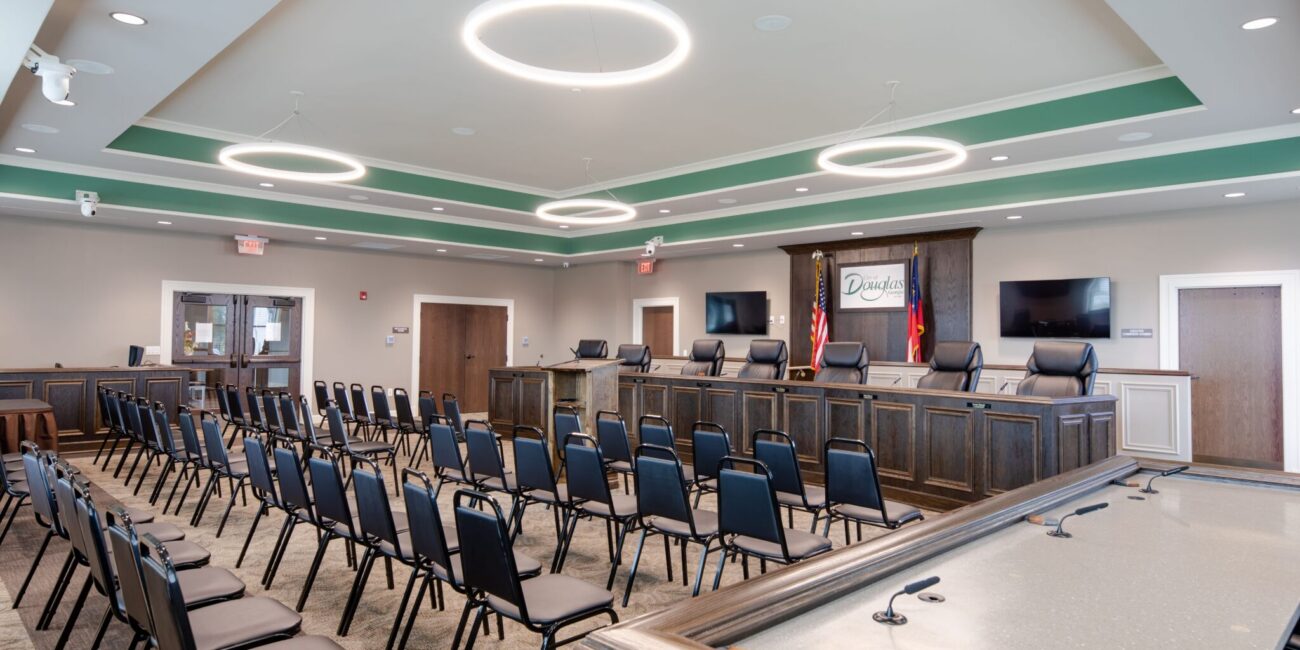 A modern, empty council chamber with rows of black chairs facing a raised panel, American flags, display screens, and a Dundas sign.