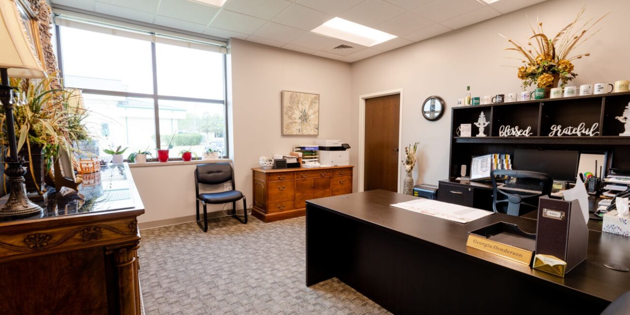 Tidy office with a large dark desk, black shelves, wooden sideboard, two chairs, window letting in light, plants and art decor.