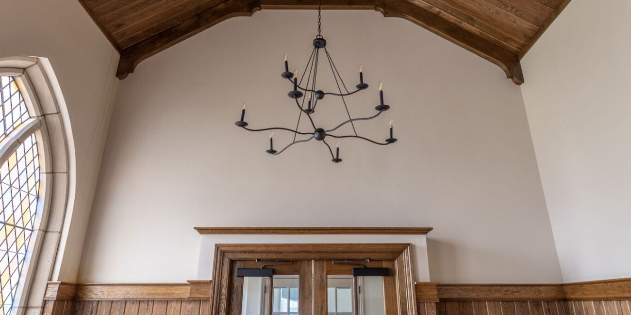 Spacious room with high wooden ceiling, black chandelier, wood-paneled walls, glass double doors, and cushioned chairs along both sides.