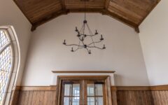 Spacious room with high wooden ceiling, black chandelier, wood-paneled walls, glass double doors, and cushioned chairs along both sides.