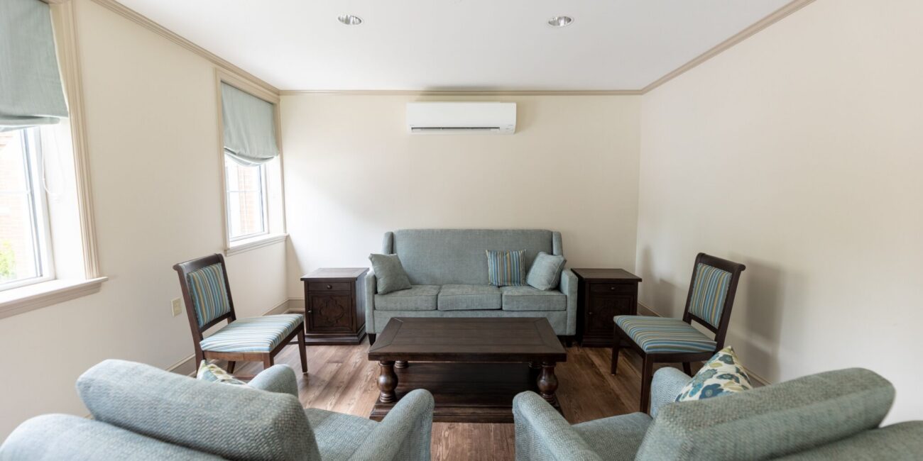 Cozy living room with light blue armchairs, sofa, wooden coffee table, side tables, blue Roman shades, and wall AC above sofa.