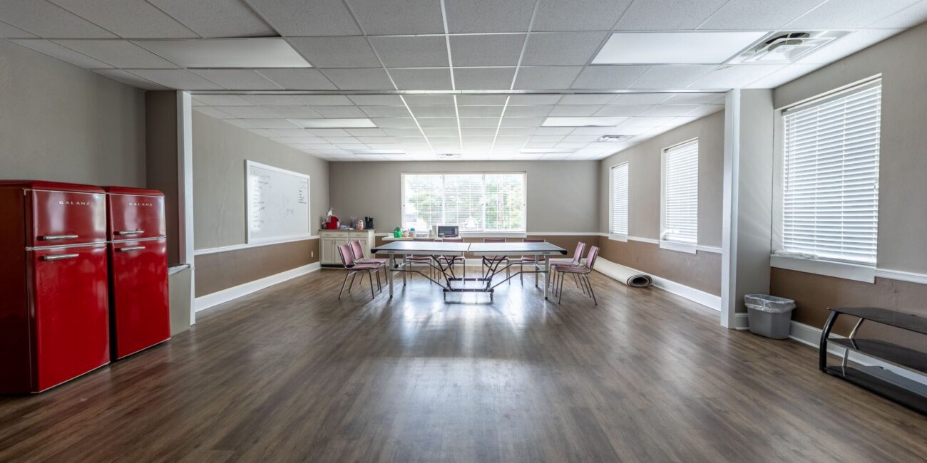 Spacious, well-lit room with wooden floors, central long table and chairs, red fridges left, large windows, whiteboard on wall.