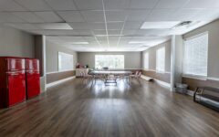 Spacious, well-lit room with wooden floors, central long table and chairs, red fridges left, large windows, whiteboard on wall.