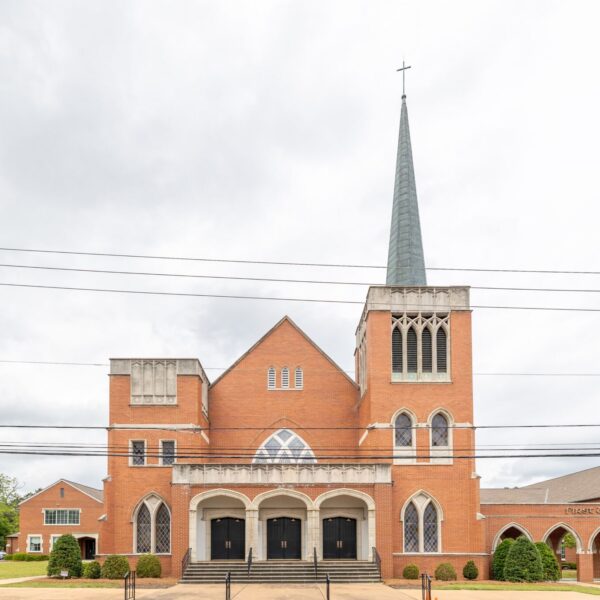 A large red-brick church with a tall steeple and cross, arched windows, manicured bushes in front, and power lines overhead.