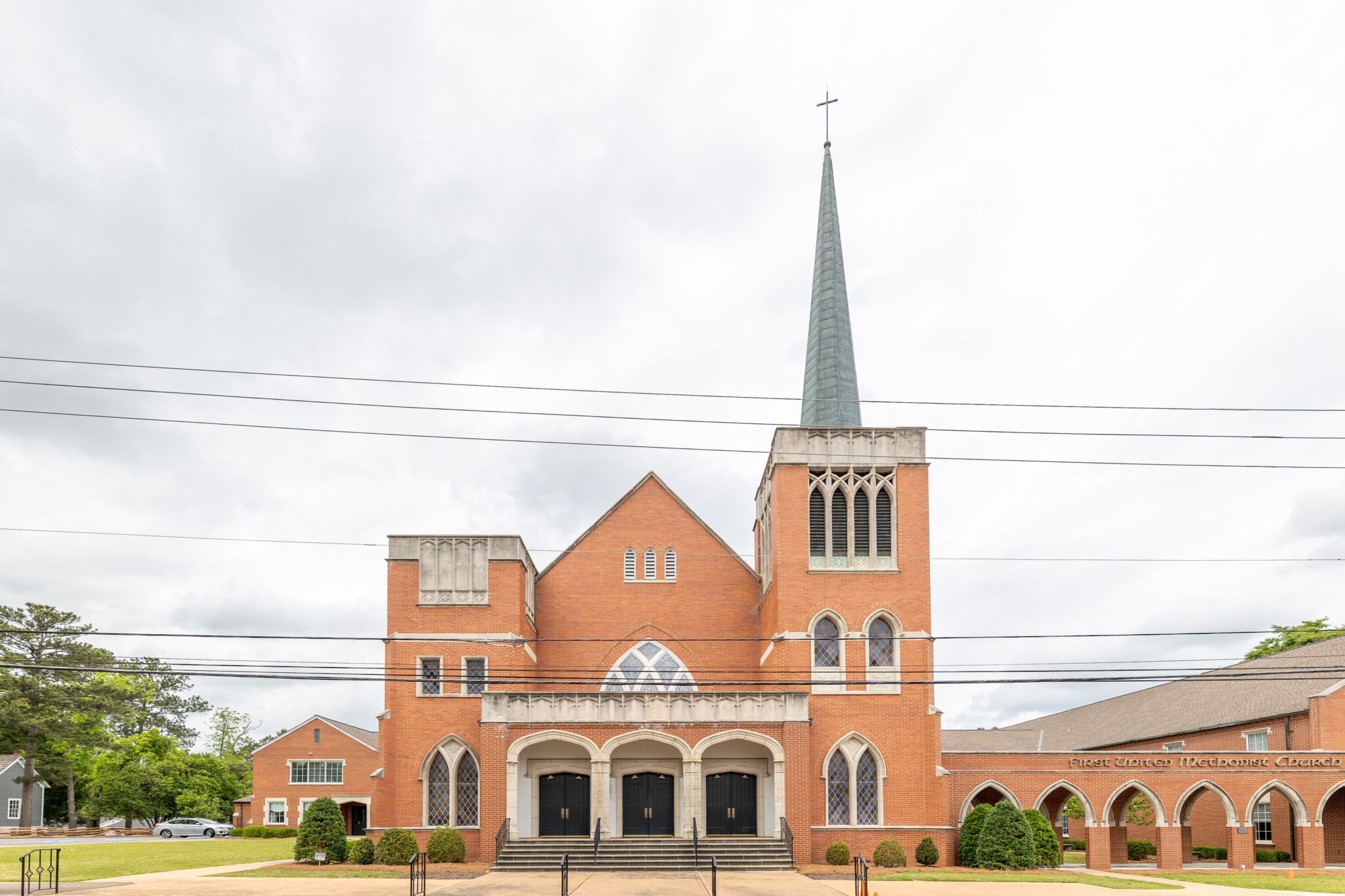 A large red-brick church with a tall steeple and cross, arched windows, manicured bushes in front, and power lines overhead.