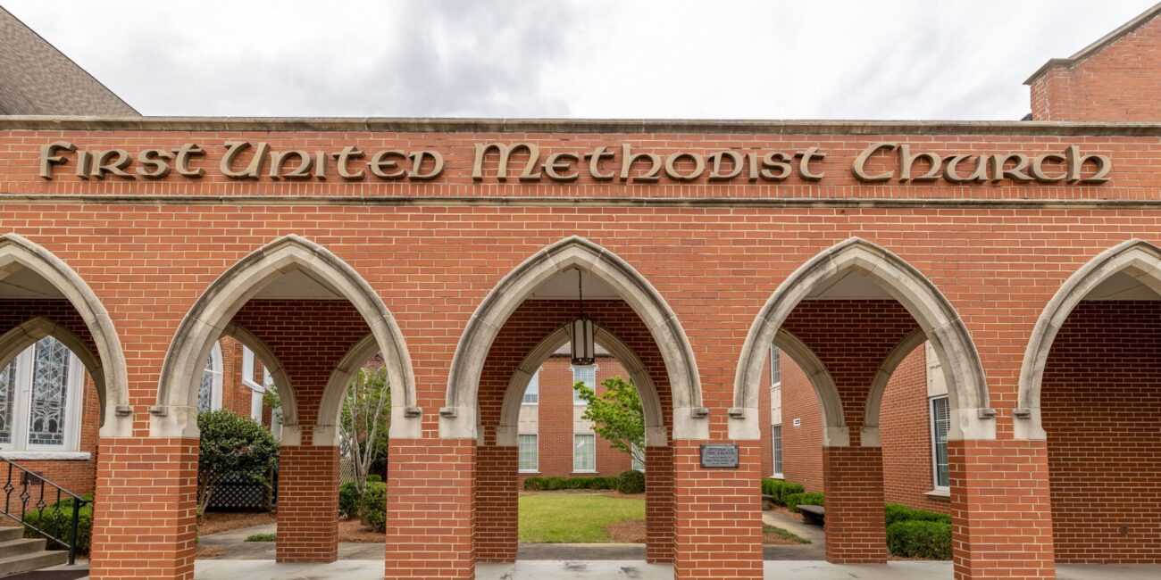 Entrance of First United Methodist Church with red brick arches, stone trim, church name above archways, and a cloudy sky behind.