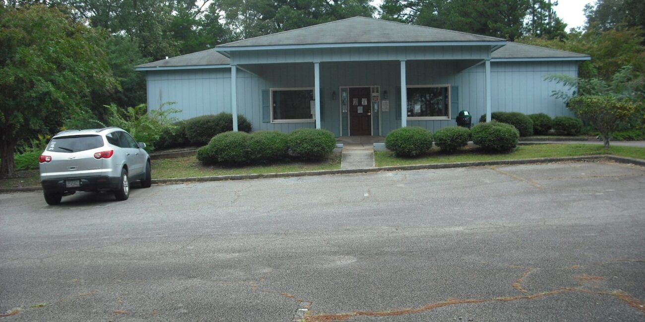 Light blue single-story building with a covered entrance, bushes and trees around, and a silver SUV in an almost empty front lot.