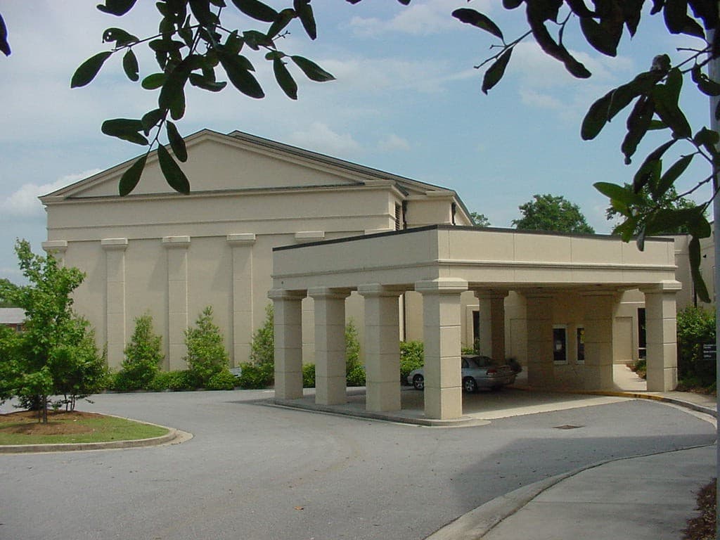 Beige building with large columns, covered drive-through entrance, small trees, car under canopy, green leaves framing top.