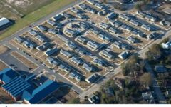 Aerial view of Felton Homes Apartments with rows of identical single-story buildings, looping streets, greenery, and nearby structures.