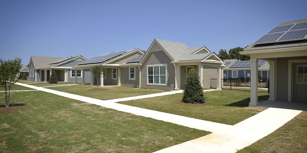 A row of one-story houses with solar-paneled roofs, tidy lawns, small trees, and crossing sidewalks under a clear blue sky.