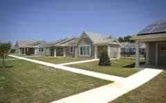 A row of one-story houses with solar-paneled roofs, tidy lawns, small trees, and crossing sidewalks under a clear blue sky.