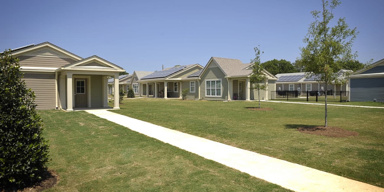 A row of single-story houses with neat lawns and young trees along a walkway under blue sky; some roofs have solar panels.