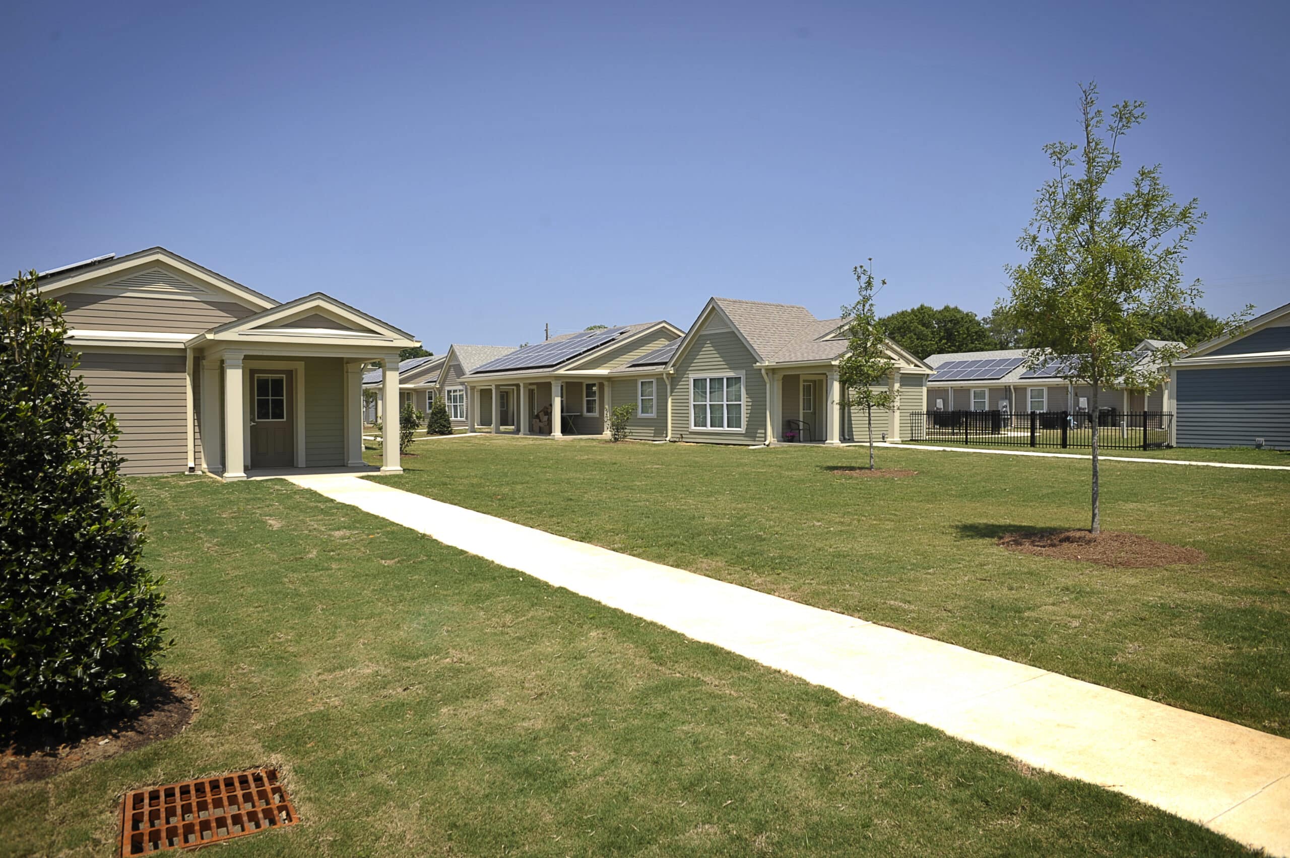 Single-story houses with porches face a grassy courtyard and central walkway under a blue sky, with young trees along the path.