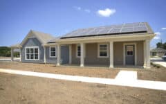 A single-story house with solar panels, gray siding, white trim, bare dirt yard, concrete walkway, porch and a sunny clear sky.