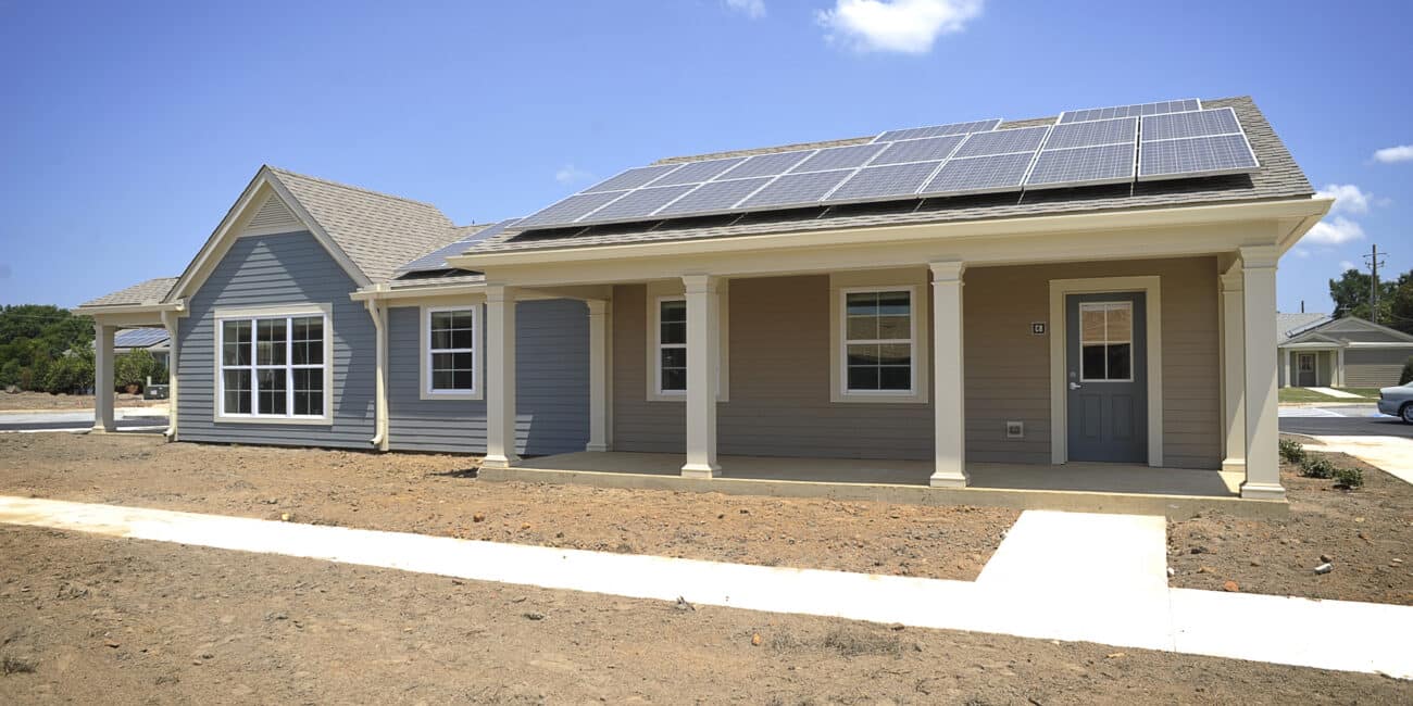 A gray single-story house with white trim and rooftop solar panels sits on a dirt yard. A concrete walkway leads to the front door.