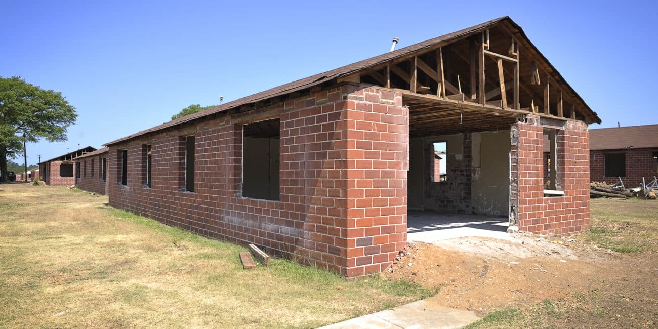 Partially demolished brick building with missing roof, exposed beams, open frames, on grass under a clear blue sky.