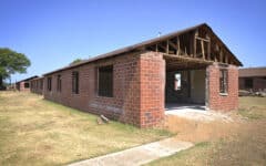 Partially demolished brick building with missing roof, exposed beams, open frames, on grass under a clear blue sky.