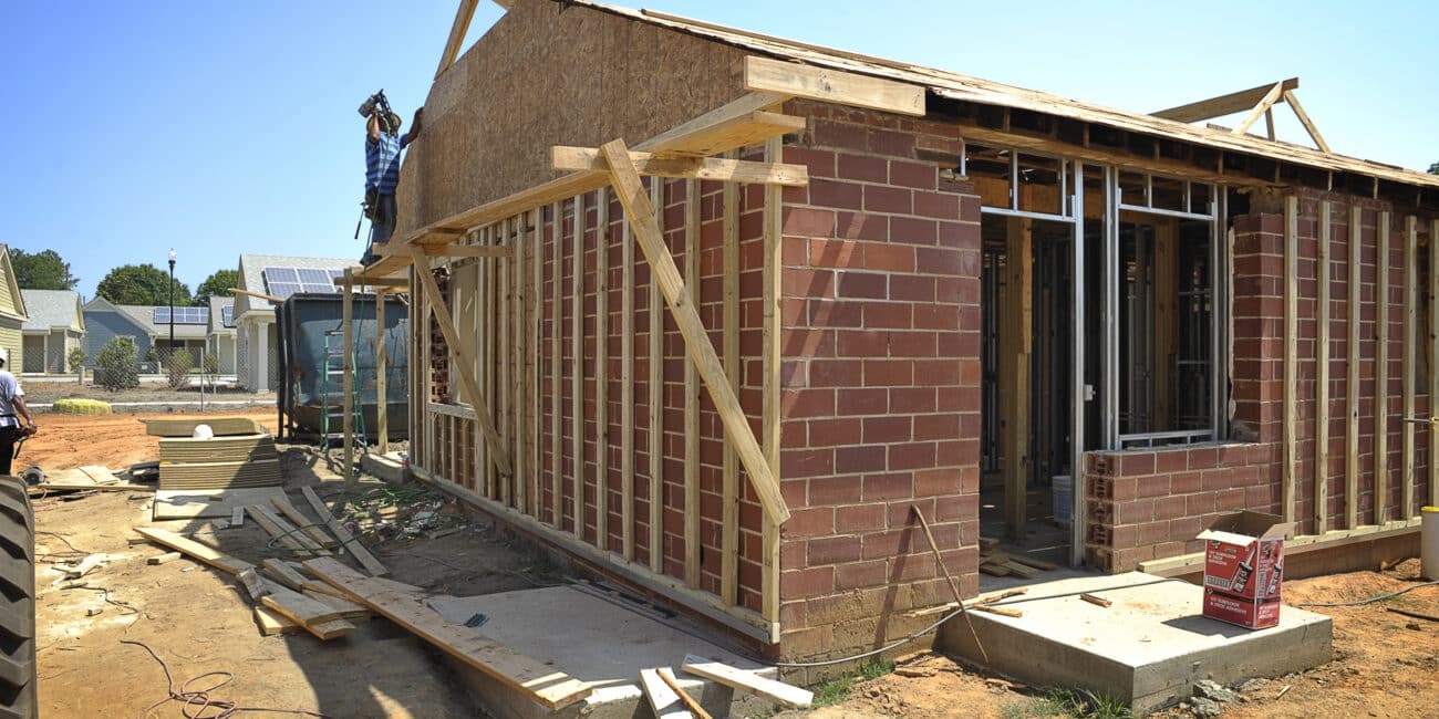 A partially built house with brick and wood framing, scattered building materials, and a worker on the roof beneath a clear blue sky.