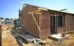 A house being built with visible wooden framing and brick walls, scattered tools, and a worker on the roof beneath a blue sky.