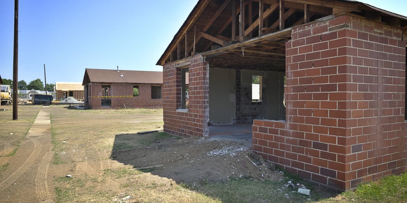 A partially demolished brick house without windows or doors stands on dry grass, with another house and equipment in the background.