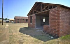 A partially demolished brick house without windows or doors stands on dry grass, with another house and equipment in the background.
