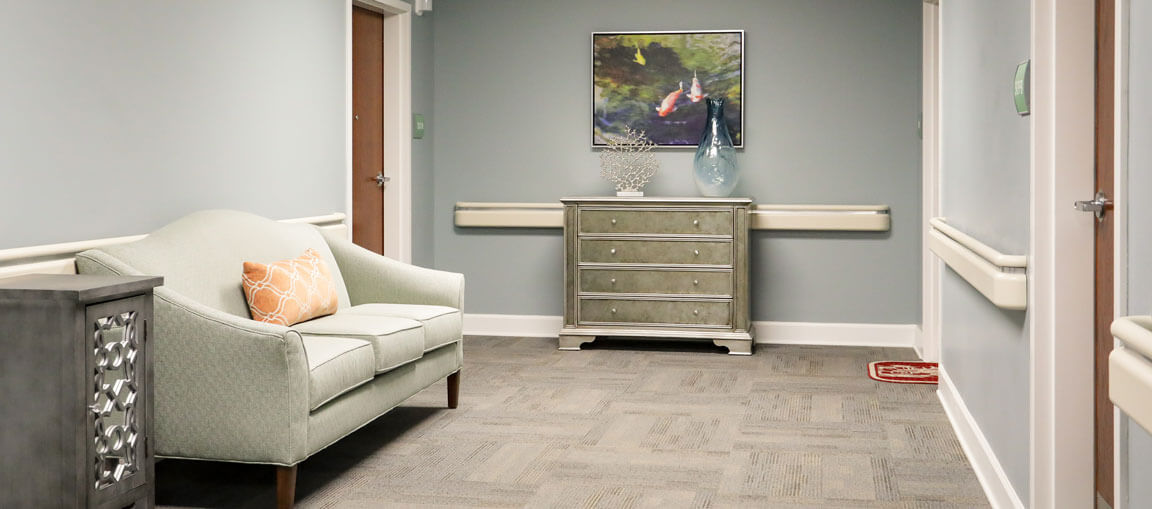 A hallway with a light green couch and orange cushion, gray drawers with vases, art on the wall, beige carpet, and modern decor.