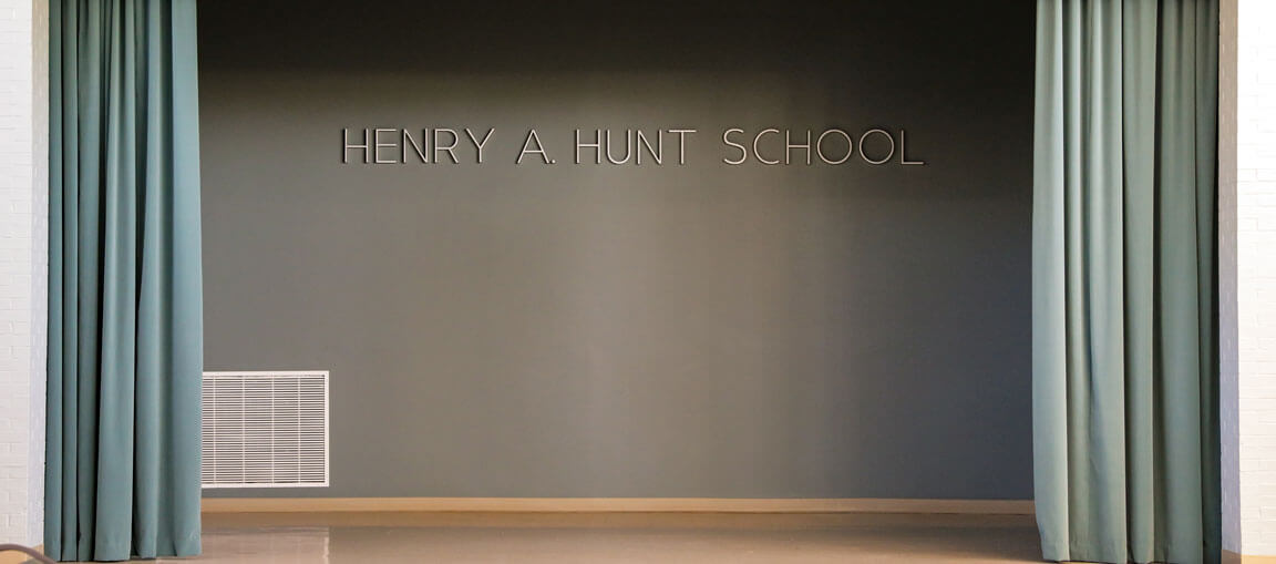 A stage with teal curtains drawn aside, revealing a gray wall with the words HENRY A. HUNT SCHOOL in large letters above a floor vent.
