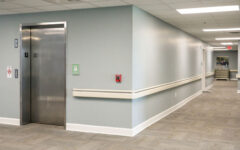 Clean, empty hallway with grey walls, beige carpet, ceiling lights, closed elevator left, handrails on walls, safety signs visible.