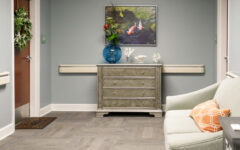 Small sitting area with light green couch, orange pillow, gray chest with blue vase and coral decor, nature painting, door with wreath.