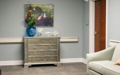 A hallway with gray walls, a silver chest topped with a blue vase and coral, a painting above, closed wood door, green couch, orange pillow.