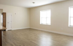 Spacious empty room with light walls, two large windows with blinds, wood-look floors, and partial kitchen counter on the left.