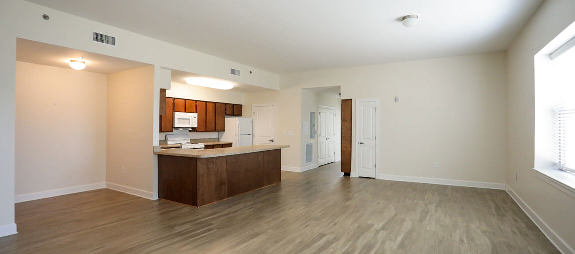 Open-plan apartment with light wood floors, white walls, big windows, brown kitchen cabinets, white appliances, and breakfast bar.