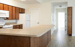 A modern kitchen with wooden cabinets, white appliances, an L-shaped countertop, light walls, wood floors, and doorways visible.