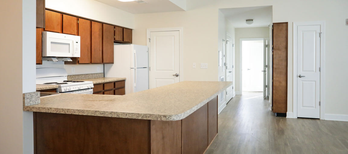 A modern kitchen with wooden cabinets, white appliances, an L-shaped countertop, light walls, wood floors, and doorways visible.