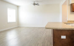 Spacious, unfurnished open-plan apartment with light wood floors, beige walls, large windows, ceiling fan, and kitchen with brown cabinets and granite countertop.