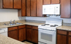 A modern kitchen features brown wooden cabinets, a white stove and oven, built-in microwave, double sink, dishwasher, and speckled light countertops.